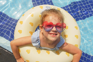 Smiling little blonde girl in pink heart-shaped glasses in the center of rubber ring in swimming pool