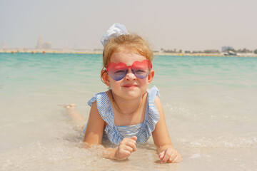 A little girl in a blue and white striped swimsuit and heart-shaped sunglasses smiles while lying in turquoise water of a beach