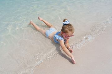 A little girl in a blue and white striped swimsuit and heart-shaped sunglasses lies on her stomach at the water's edge, playfully reaching for the sand as gentle waves lap around her on a sunny beach