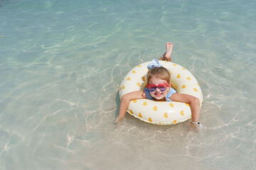 A joyful little girl with heart-shaped sunglasses smiles brightly while floating on a yellow heart-patterned inflatable ring in the clear, sun-dappled turquoise water of a tropical beach