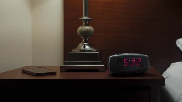 In a dark room, an alarm clock glows on a nightstand. A person turns on the light and shuts off the alarm, ideal for morning, sleep, and lifestyle visuals.