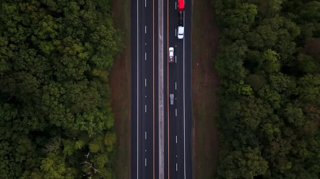 A top down view over a highway with two lanes in each direction with traffic only in one direction. The camera is tilted down and stationary on a cloudy day. Green trees on either side of the roadway.