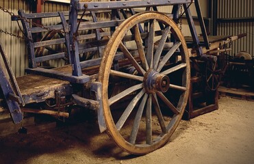A weathered blue cart rests quietly in the shadows of a corrugated shed, its spoked wheel standing like a relic of forgotten journeys. Chains and wood speak of labor and time