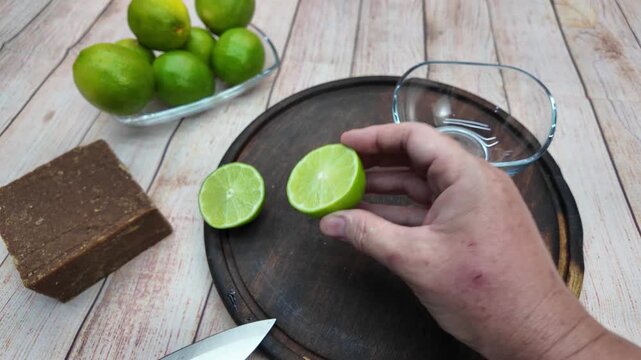 Close-up shot showing human hands slicing a fresh green lime with a knife on a rustic wooden cutting board, next to a block of panela and bowl.