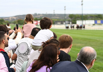 Espectadores y p&uacute;blico con adultos y ni&ntilde;os a hombros en caballito animando y personas atentas a carrera de caballos en pista de hierba del hip&oacute;dromo