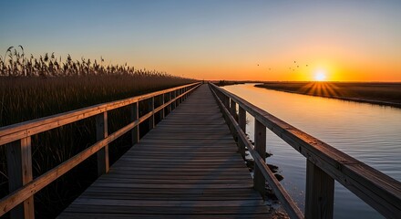 Obraz premium Wooden boardwalk stretching into the distance alongside a tranquil river at sunset, with golden light reflecting on the water and reeds lining the banks.