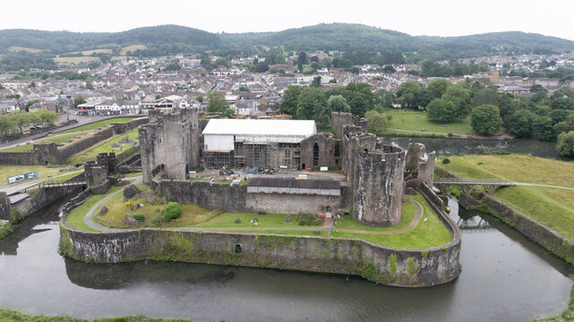 Aerial view of the imposing Caerphilly Castle, its weathered stone walls embraced by a serene moat reflecting the overcast sky, a testament to centuries of history, Caerphilly, Wales, United Kingdom.