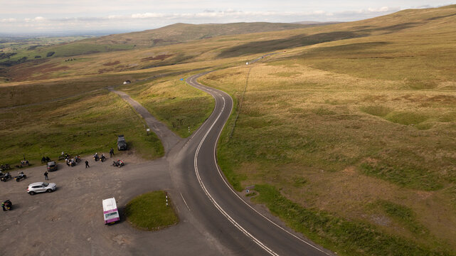 Aerial view of the winding road cutting through the golden-brown moorland, contrasting with the gathering of vehicles at the roadside rest stop, Hartside Pass, Alston, England, United Kingdom.