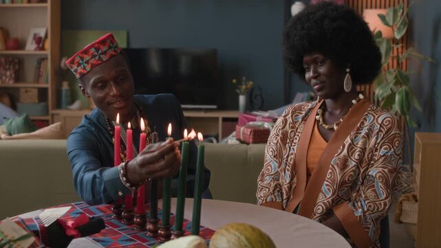Waist up of Black man and woman sharing Kwanzaa traditions, lighting wax candles on red patterned table in their cozy living room, celebrating family, culture, and togetherness