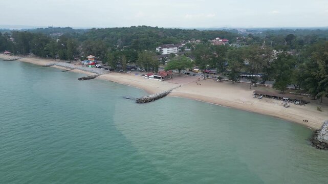 A beach with a rocky shoreline and a few buildings in the distance. The water is calm and blue. Aerial Tankong Kling, Melaka
