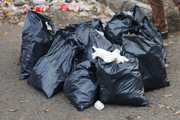 A pile of black garbage bags sits on a street, with scattered trash and disposable cups in the background. The scene reflects urban waste, pollution, and sanitation issues.