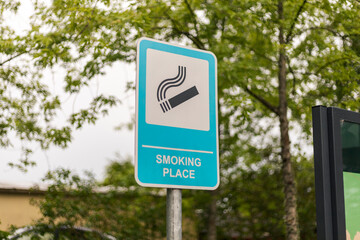 A sign indicating a designated smoking area is visible in a green park setting. Surrounding trees add to the natural ambiance of the spot for smokers