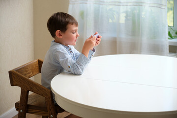 Young charming boy sitting at wooden table and holding a phone in his hands. Concentrated kid playing mobile game or watching video. Child using social media. A child aged 4 years (four years old)