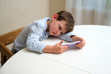 Naklejka premium Little boy, a child, laying arm on table intently looking at a cell phone screen while engaging with educational games or cartoons for development. A child aged 4 years (four years old)
