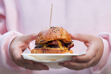 A close-up of young woman carrying a gourmet burger  with melted cheese on a tray with napkins at Karlin food festival in Prague.