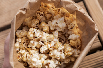 A hand holds a brown paper bag filled with fluffy, golden popcorn. The warm sunlight illuminates the popcorn as it sits on a wooden surface, perfect for snacking at a gathering