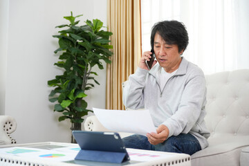 Mature Man Working Remotely, Talking on Smartphone and Reviewing Documents at Home, Middle-Aged Asian Businessman Conducting a Video Conference Call while Looking at Paperwork