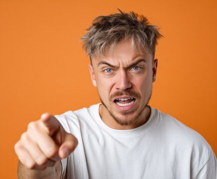 A man with a distressed expression, wearing a white t-shirt, directs an accusatory finger towards the viewer against a vibrant orange backdrop.
