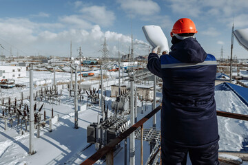 Worker on an elevated platform oversees a vast, snow-covered power substation, emphasizing energy management, industrial scale, and critical infrastructure. Ideal for utilities and cold weather