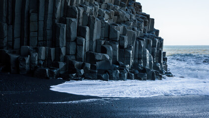 A basalt column cliff meets the ocean waves on a black sand beach on a cloudy day outside nature