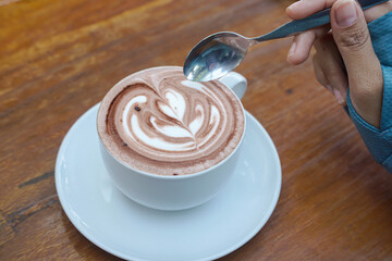 Cappuccino coffee with heart-shaped latte art on a wooden table, a close-up photo of a cappuccino drink in a white cup with a spoon on a wood background.