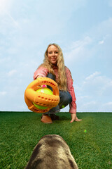 Joyful woman leaning forward with ball launcher as seen from playful dog on grass. Concept of...