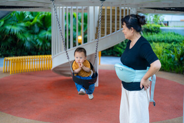 Grandma takes her grandson to the playground. The toddler swings on the swing, wearing an orange short-sleeved shirt. The one-year-old baby. The retirement lifestyle of Chinese seniors. Families with 