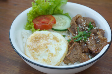A vibrant bowl of rice topped with grilled beef, a fried egg, and colorful vegetables, served in a rustic dish