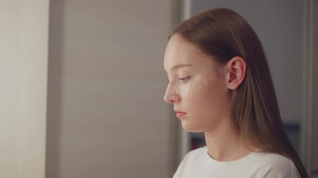 Caucasian young woman in white t-shirt applying concealer on her face indoors, side view, focused expression, symbolizing skincare, makeup process and beauty routine.