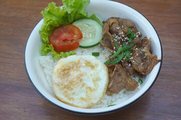 A vibrant bowl of rice topped with grilled beef, a fried egg, and colorful vegetables, served in a rustic dish