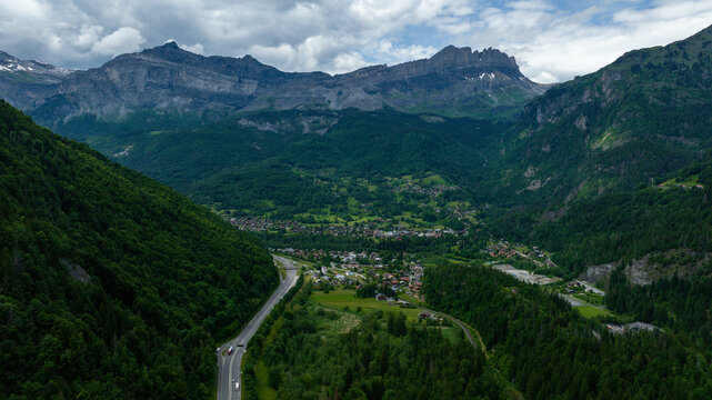 Aerial view of a winding road cutting through a verdant valley, nestled between towering, jagged mountains under a cloudy sky, Chamonix, France.