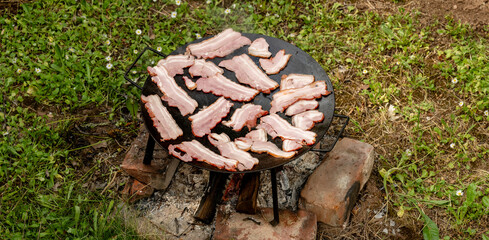 Cooking strips of raw bacon on a cast iron griddle over an open fire outdoors. Rustic, outdoor cooking setup in a natural, grassy environment with firewood flames beneath the pan.