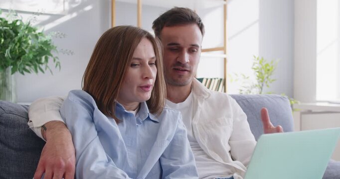 Loving young family couple relaxing on sofa with laptop, hugging and enjoying conversation. focused wife listening to her husband, who gesturing while explaining and sharing something interesting.