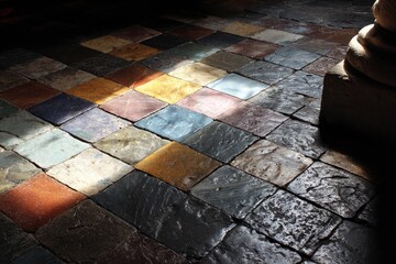 Sunlit multicolored tile floor, varied hues and textures, showing wear, near a stone column in shadow