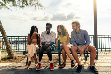 Happy diverse friends laughing and sharing a smartphone on a summer day. Multicultural group enjoying togetherness on a park bench.