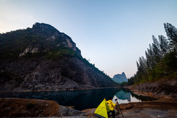 A serene camping scene with a bright green tent and a traveler by a calm mountain lake at dawn, surrounded by rugged cliffs and pine trees reflecting on the water.