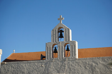 Campanile bianco con 3 campane e una croce, con tetto rosso sullo sfondo. Particolare del monastero di Odigitria sud isola di Creta