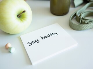 A green apple sits next to a note saying stay healthy. Colorful vitamins and a strap from a bag complete the scene on a clean white table, promoting wellness in daily life.