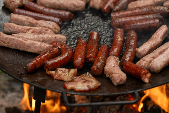 A large, round, flat metal grill, discada or plow disc grill, being used over an open flame. Sausages, pork and beef steaks, and kebabs are cooked on it. A little greenery is visible in the background