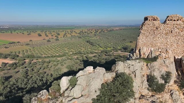 Low-angle drone flight in 70mm over the ruins of Dos Hermanas Castle in Navahermosa, Toledo. The shot glides near the ancient stone walls on the rocky outcrop, surrounded by olive groves and forests.