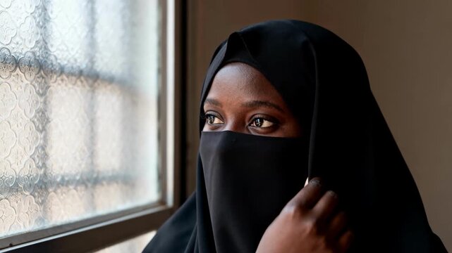 African woman wearing black niqab looking out a patterned glass window in soft daylight, quiet indoor portrait with thoughtful expression and cultural clothing detail for editorial and social themes