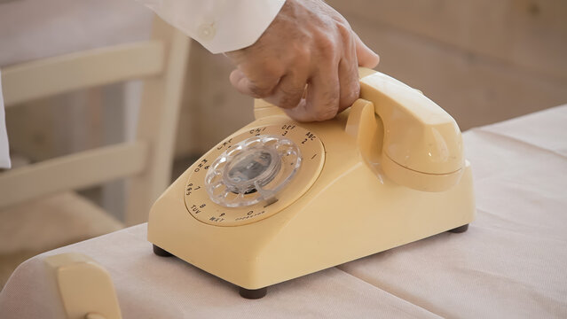 Hand dialing vintage yellow rotary telephone on white table in soft neutral interior. Close-up captures nostalgic communication device with clear rotary dial detail. Retro aesthetic perfect for timele - Powered by Adobe