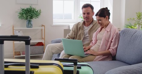 Discussion of smiling couple planning travel holidays online, using laptop together at home. Young happy tourists discuss plan of trip, sitting on sofa in living room, suitcases ready for vacation - Powered by Adobe