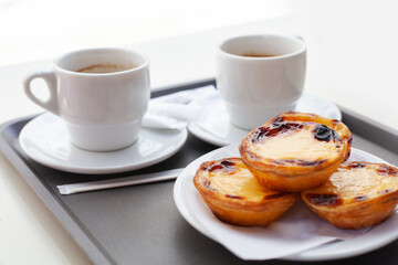 Close-up of traditional Portuguese Pastel de Nata on a plate with mugs of black coffee. Crispy custard tarts and espresso on rustic table, perfect for breakfast or dessert