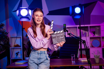 Cute Young female podcaster holding clapperboard in vibrant studio setup with neon lighting
