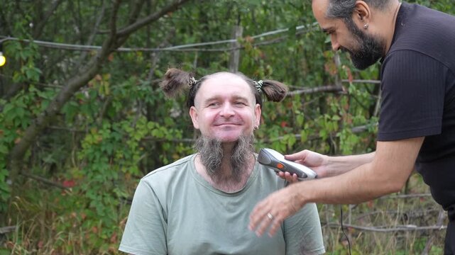 A bearded man with comical hair braids is having his beard shaved with an electric trimmer outdoors, humorous makeover scene in the garden, funny haircut transformation