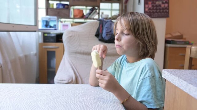 Young boy sitting at a kitchen table holding a vanilla ice cream bar, enjoying a frozen treat at home with potted plants in the background