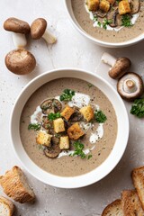 Overhead photo of a bowl of creamy mushroom soup with toasted croutons, white sauce drizzle, and green herbs surrounded by shiitake mushrooms and bread slices