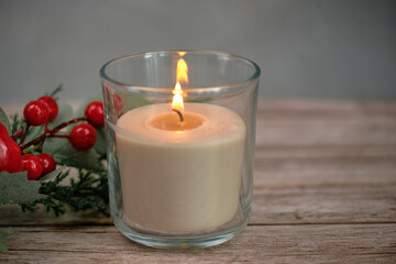 Close-up of a burning candle in a glass glass on a wooden table. Next to it is a twig with bright red berries, creating a festive New Year's mood.