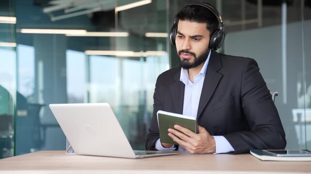 Businessman in wireless headphones watching video call conference takes notes looking at laptop screen sitting in business office. Worker in suit listening to remote online course, seminar or training